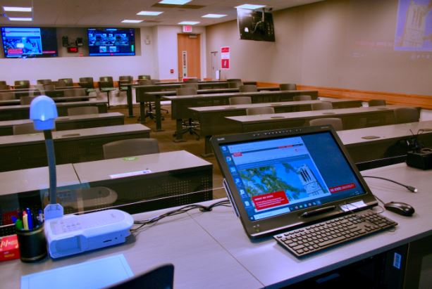 View from the front of a DELTA classroom showing the instructor's view of the screen and controls at the front of the room, and large monitors at the back of the room showing the shared screen content and gallery view of Zoom participants.
