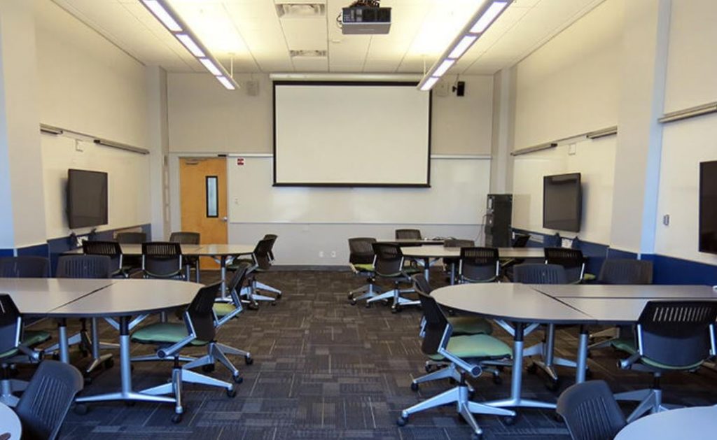 An active learning classroom at UC Berkeley containing groups of chairs around tables. Large wall monitors next to each table allow for interaction with online attendees.